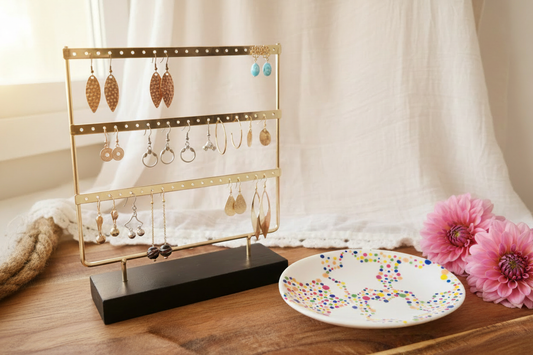 Gouden oorbellenrek met houten voet en handgeschilderd sieradenschaaltje met kleurrijke stippen, gestileerd met oorbellen collectie, witte linnen en roze dahlia bloemen